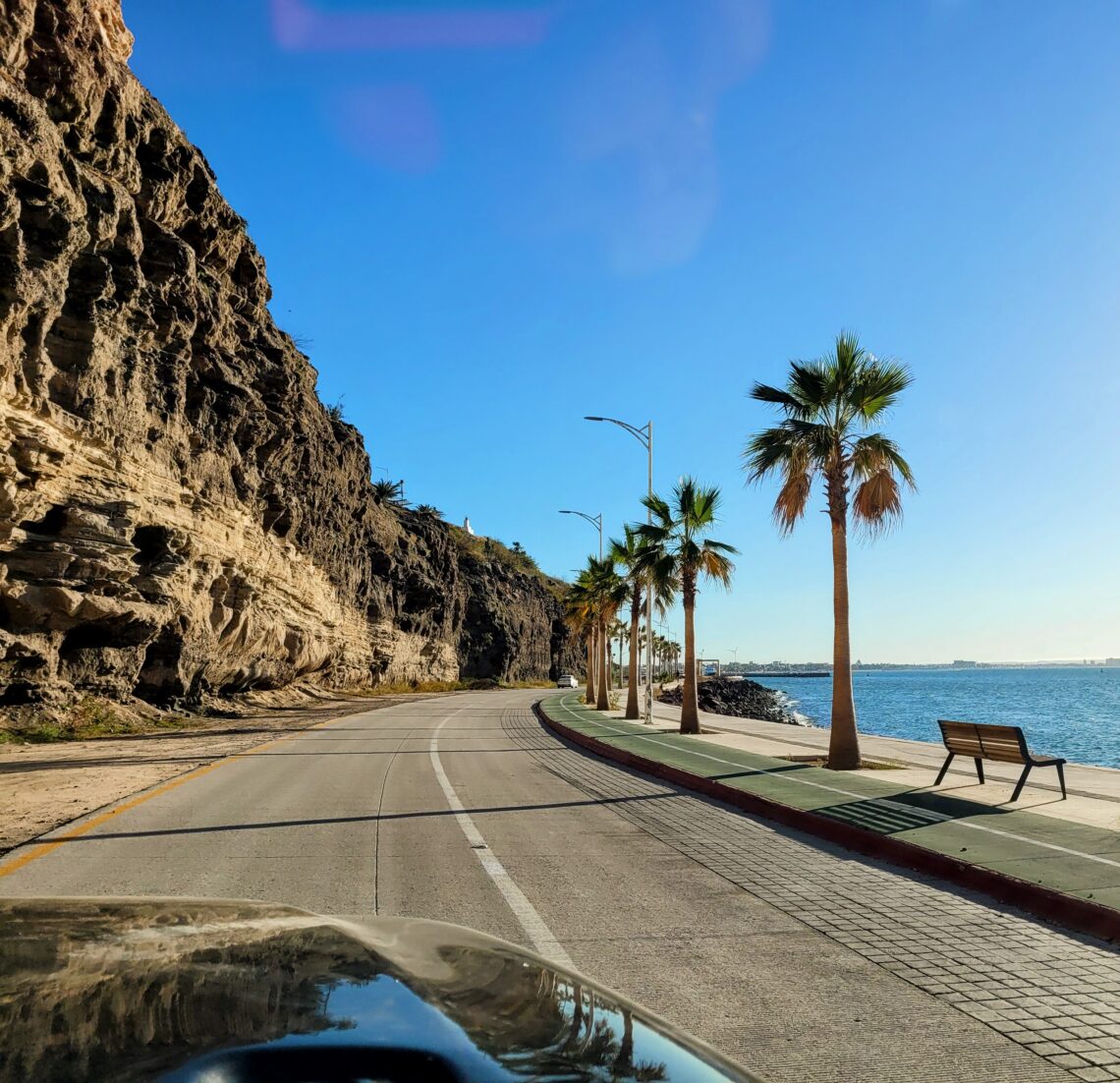 A Jeep drive along the coast with a clear blue sky. There's a rock cliffside to the left and the sea side is lined with palm trees; La Paz, Baja California Sur (Mexico)