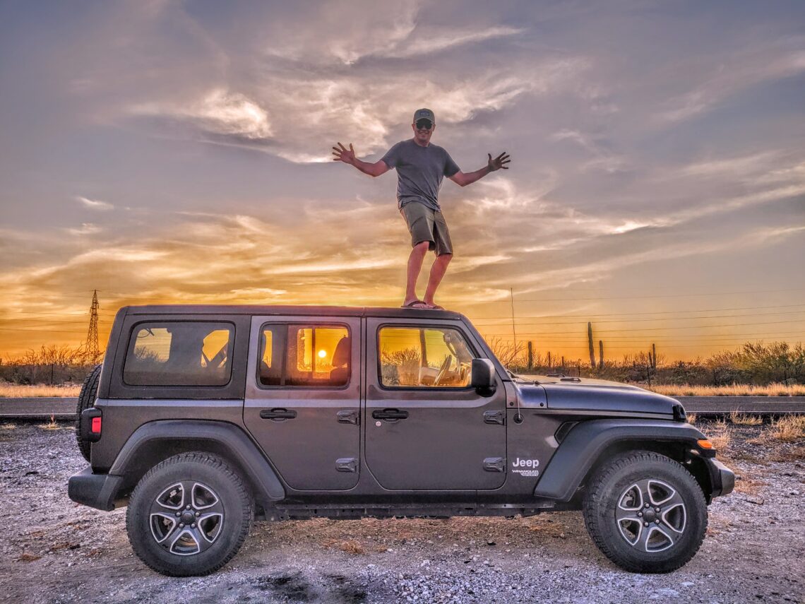 Paul is standing on top of a black Jeep Wrangler with a gorgeous sunset lighting up the sky behind him and through the Jeep's windows; Baja California Sur, Mexico