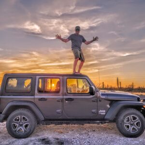 Paul is standing on top of a black Jeep Wrangler with a gorgeous sunset lighting up the sky behind him and through the Jeep's windows; Baja California Sur, Mexico