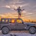 Paul is standing on top of a black Jeep Wrangler with a gorgeous sunset lighting up the sky behind him and through the Jeep's windows; Baja California Sur, Mexico