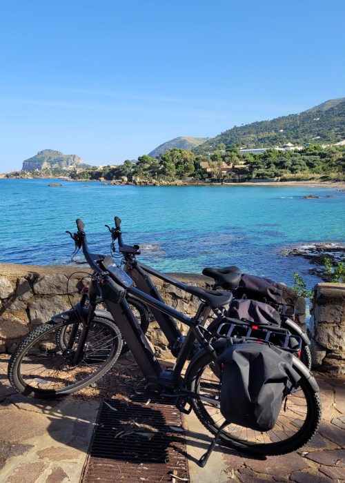 Two e-bikes against a rock wall with Spiaggia di Mazzaforno in the background; Sicily, Italy
