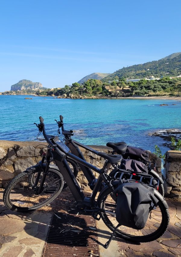 Two e-bikes against a rock wall with Spiaggia di Mazzaforno in the background; Sicily, Italy