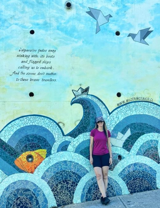 Large blue and white mural with splashing waves and birds flying and me standing in front with a poem that says "Valparaiso fades away, winking with its boots, and flagged ships, calling us to embark. And the sirens don't matter to these brave travellers"; Valparaíso, Chile