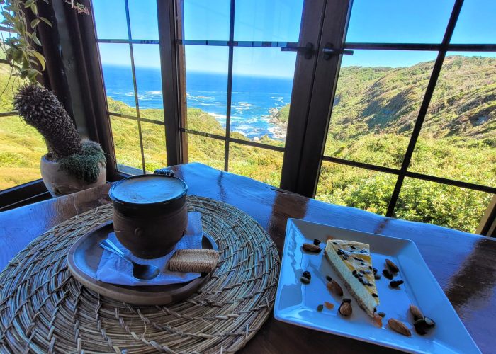 Ocean view through a large window with a cappuccino and a slice of passionfruit cheesecake at Explore - Café & Lounge; Chiloé Island, Chile